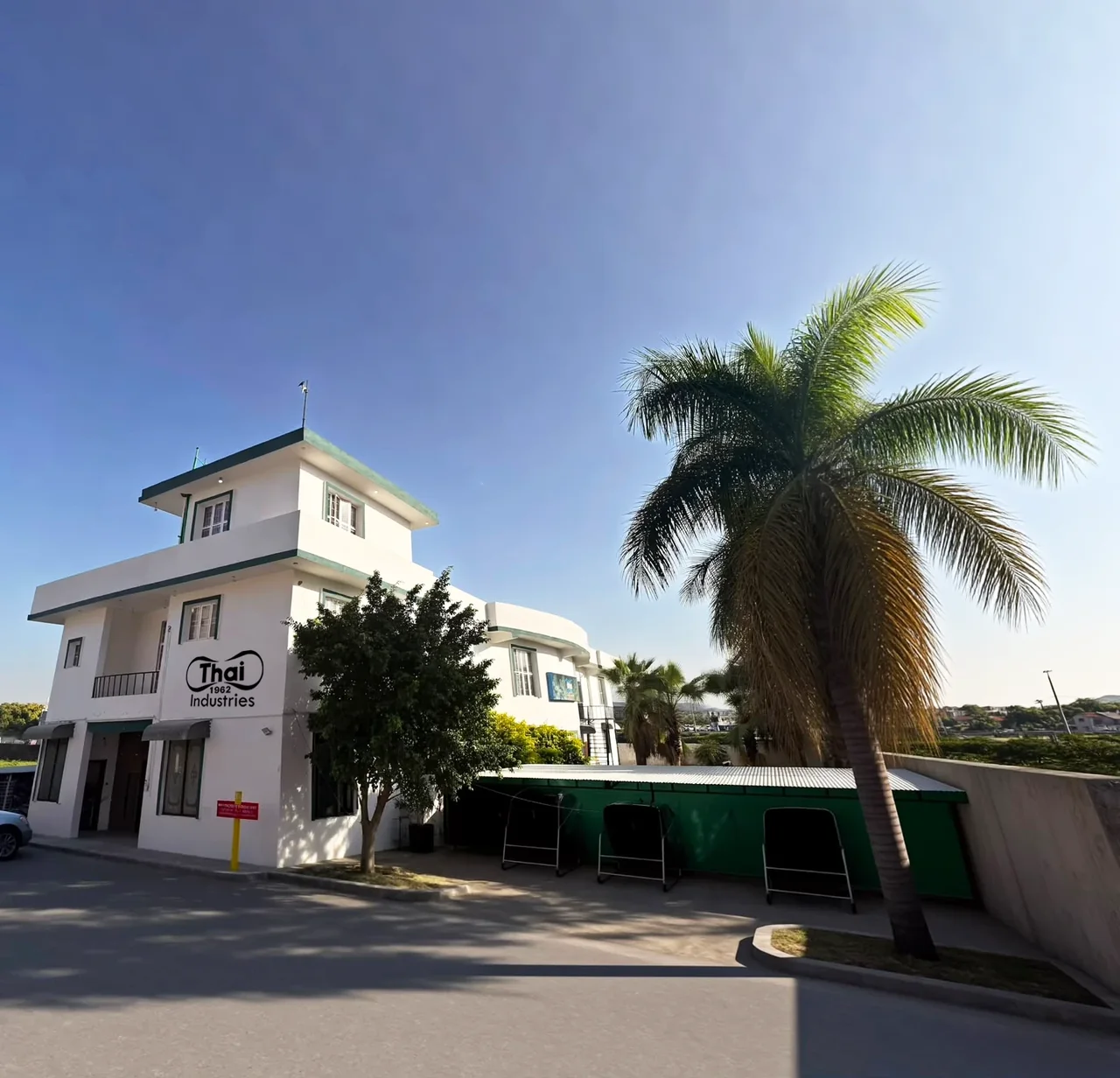 The Thai Industries factory seen from the access road: a white multi-storey building with the Thai Industries 1962 logo on its wall, a covered parking bay and a tall palm tree